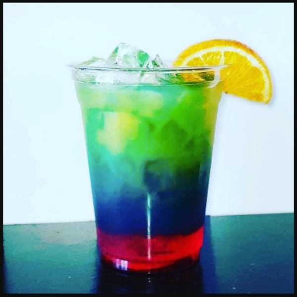 A vibrant, layered rainbow drink with ice and an orange slice in a clear plastic cup.