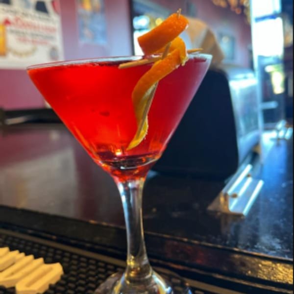 A red cocktail in a martini glass, garnished with an orange peel, on a dark bar.